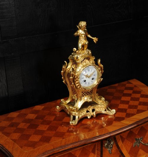 fig 4. A French Rococo gilt bronze mantel clock featuring a cupid figure at the top, scrolling foliage, and a white enamel dial with Roman numerals.