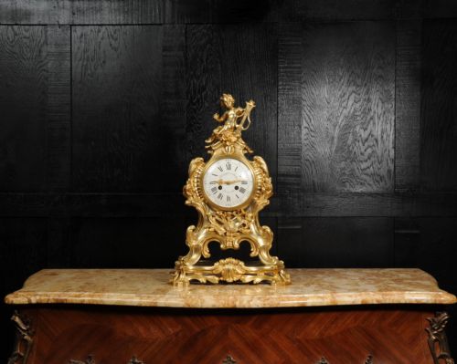 fig 1. A French Rococo gilded bronze mantel clock featuring a cherub surmount, musical instrument reliefs, and a white enamel dial.