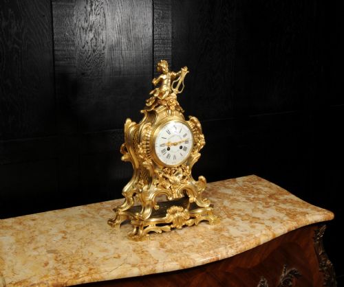 fig 3. A French Rococo gilded bronze mantel clock featuring a cherub surmount, musical instrument reliefs, and a white enamel dial.