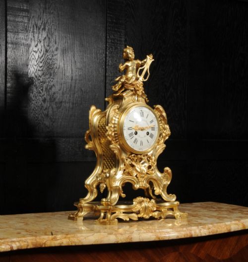 fig 5. A French Rococo gilded bronze mantel clock featuring a cherub surmount, musical instrument reliefs, and a white enamel dial.