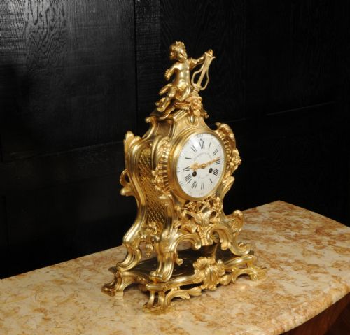 fig 6. A French Rococo gilded bronze mantel clock featuring a cherub surmount, musical instrument reliefs, and a white enamel dial.