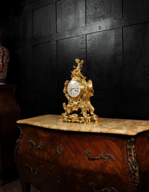 fig 7. A French Rococo gilded bronze mantel clock featuring a cherub surmount, musical instrument reliefs, and a white enamel dial.