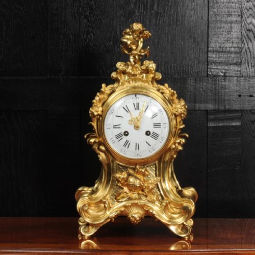 fig 1. A French Rococo gilded bronze mantel clock featuring an ornate scrollwork case, a Cupid finial, and a white enamel dial with Roman numerals.