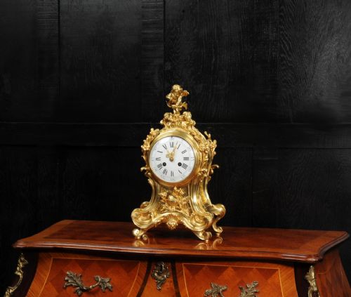 fig 2. A French Rococo gilded bronze mantel clock featuring an ornate scrollwork case, a Cupid finial, and a white enamel dial with Roman numerals.