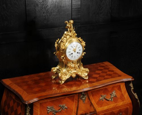 fig 3. A French Rococo gilded bronze mantel clock featuring an ornate scrollwork case, a Cupid finial, and a white enamel dial with Roman numerals.