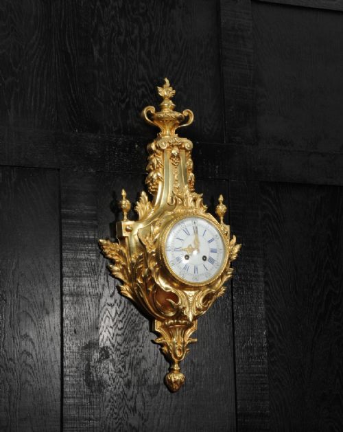 fig 3. A Louis XVI style gilt bronze cartel wall clock featuring an urn finial, detailed acanthus leaf scrolls, and a white enamel dial with Roman numerals.