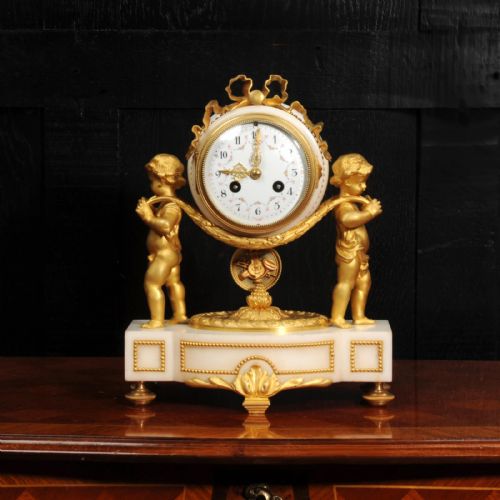 fig 1. An antique French mantel clock crafted from white marble and gilt bronze, featuring two sculpted cherubs framing an enamelled circular dial topped with a ribbon crest.