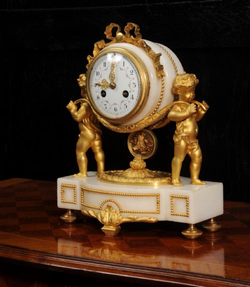 fig 2. An antique French mantel clock crafted from white marble and gilt bronze, featuring two sculpted cherubs framing an enamelled circular dial topped with a ribbon crest.