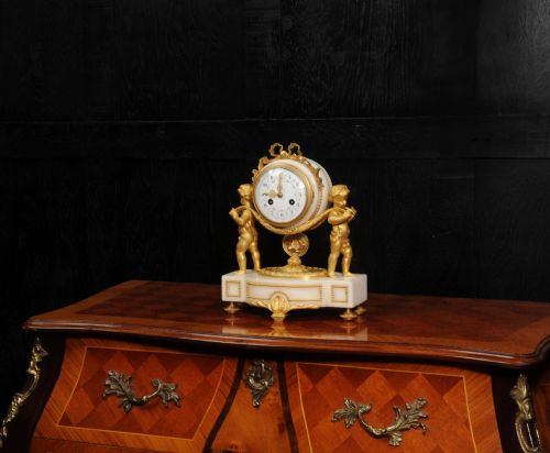 fig 3. An antique French mantel clock crafted from white marble and gilt bronze, featuring two sculpted cherubs framing an enamelled circular dial topped with a ribbon crest.