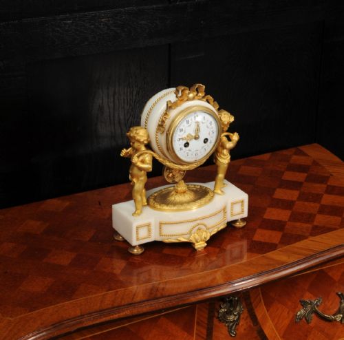 fig 4. An antique French mantel clock crafted from white marble and gilt bronze, featuring two sculpted cherubs framing an enamelled circular dial topped with a ribbon crest.