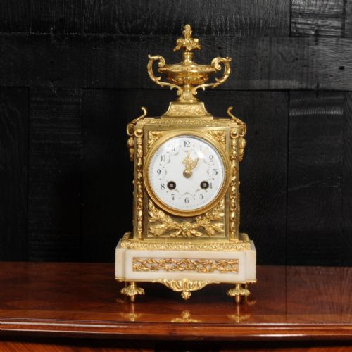 fig 1. An antique French Louis XVI style ormolu mantel clock featuring a top urn finial, a white enamel dial with floral decoration, and a white marble base with gilt metal mounts.