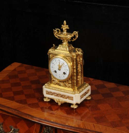 fig 2. An antique French Louis XVI style ormolu mantel clock featuring a top urn finial, a white enamel dial with floral decoration, and a white marble base with gilt metal mounts.