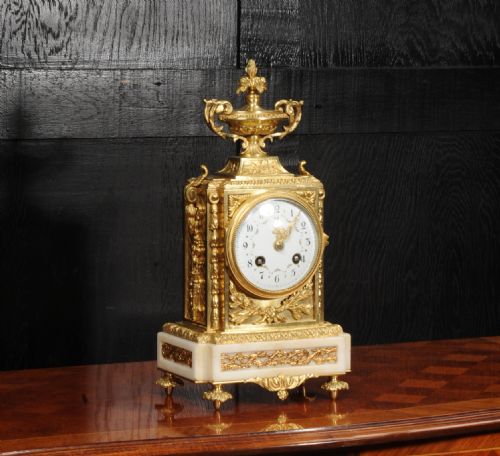 fig 3. An antique French Louis XVI style ormolu mantel clock featuring a top urn finial, a white enamel dial with floral decoration, and a white marble base with gilt metal mounts.