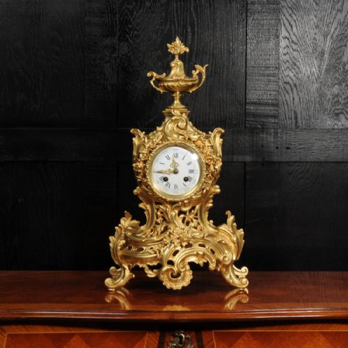 fig 1. An antique French Rococo gilt bronze mantel clock featuring scrollwork, an urn finial, and a white enamel dial with Roman numerals.