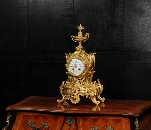 fig 2. An antique French Rococo gilt bronze mantel clock featuring scrollwork, an urn finial, and a white enamel dial with Roman numerals.