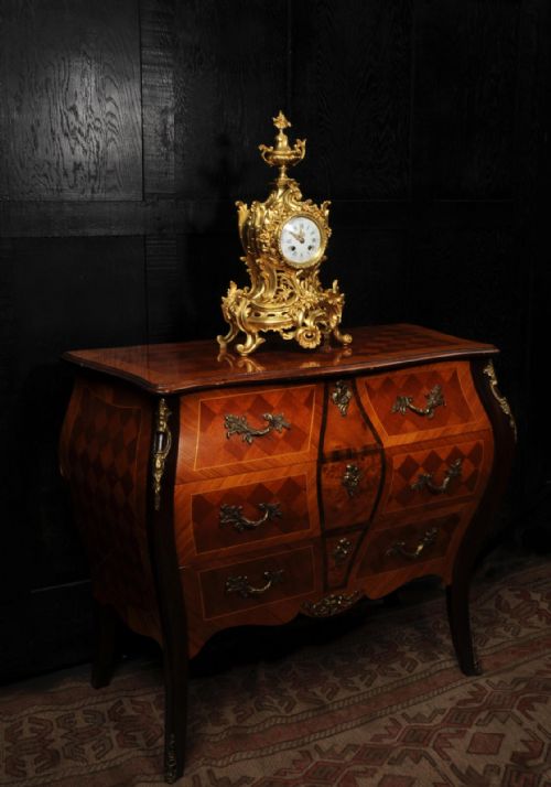 fig 3. An antique French Rococo gilt bronze mantel clock featuring scrollwork, an urn finial, and a white enamel dial with Roman numerals.