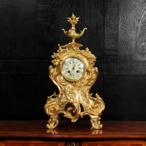 fig 1. An ornate gilt bronze Rococo mantel clock featuring scrollwork, floral carvings, a top urn finial, and a white enamel dial with Roman numerals.