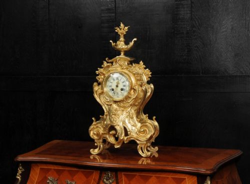 fig 2. An ornate gilt bronze Rococo mantel clock featuring scrollwork, floral carvings, a top urn finial, and a white enamel dial with Roman numerals.