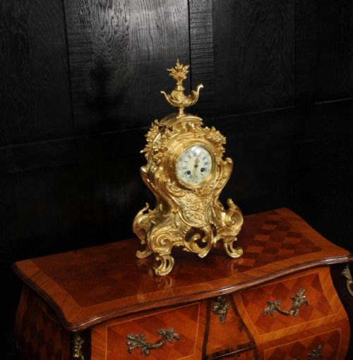 fig 3. An ornate gilt bronze Rococo mantel clock featuring scrollwork, floral carvings, a top urn finial, and a white enamel dial with Roman numerals.