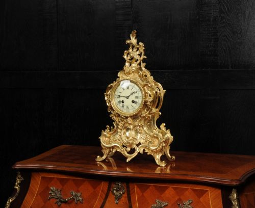 fig 1. An ornate gold gilt bronze mantel clock in the Rococo style featuring asymmetric scrollwork, acanthus leaf motifs, and a white enamel dial with black Roman numerals.