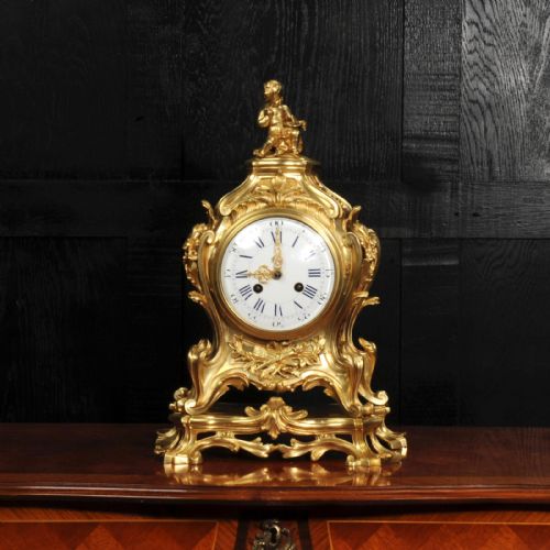 fig 1. An antique French gilt bronze Rococo mantel clock featuring an ornate scrollwork case topped with a seated cherub figure and a white enamel dial.