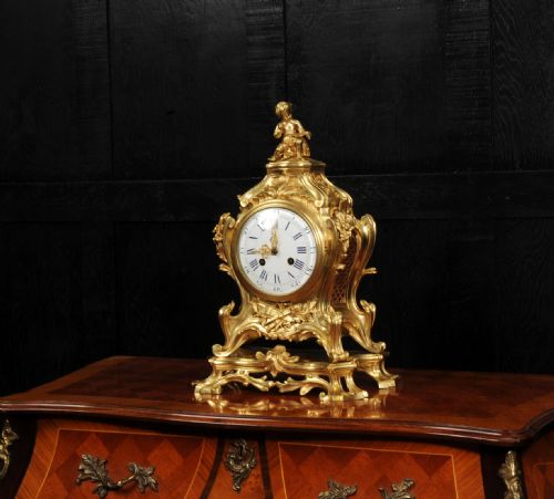 fig 2. An antique French gilt bronze Rococo mantel clock featuring an ornate scrollwork case topped with a seated cherub figure and a white enamel dial.