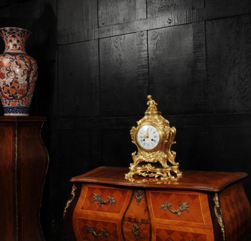 fig 3. An antique French gilt bronze Rococo mantel clock featuring an ornate scrollwork case topped with a seated cherub figure and a white enamel dial.