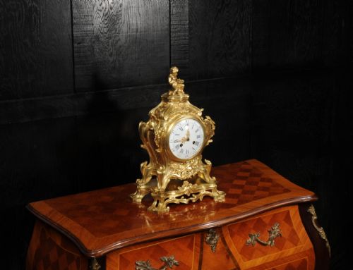 fig 4. An antique French gilt bronze Rococo mantel clock featuring an ornate scrollwork case topped with a seated cherub figure and a white enamel dial.