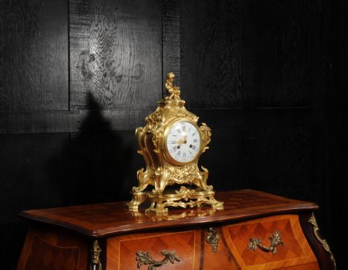 fig 5. An antique French gilt bronze Rococo mantel clock featuring an ornate scrollwork case topped with a seated cherub figure and a white enamel dial.