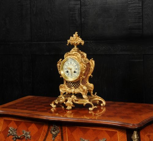 fig 1. An ornate French gilt bronze Rococo mantel clock featuring elaborate scrollwork casing, an urn finial, pierced lattice sides, and an enamelled clock face with Roman numerals.