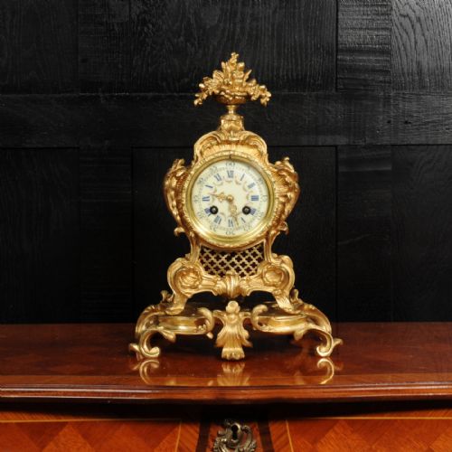 fig 2. An ornate French gilt bronze Rococo mantel clock featuring elaborate scrollwork casing, an urn finial, pierced lattice sides, and an enamelled clock face with Roman numerals.