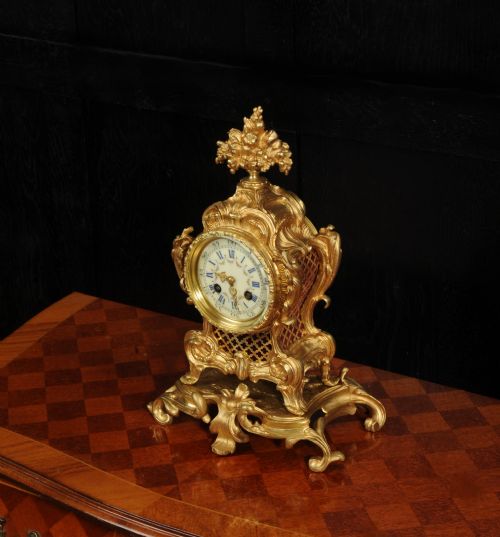 fig 3. An ornate French gilt bronze Rococo mantel clock featuring elaborate scrollwork casing, an urn finial, pierced lattice sides, and an enamelled clock face with Roman numerals.