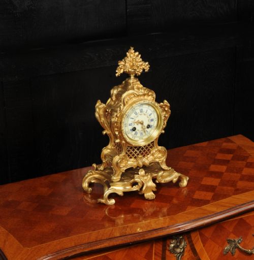 fig 4. An ornate French gilt bronze Rococo mantel clock featuring elaborate scrollwork casing, an urn finial, pierced lattice sides, and an enamelled clock face with Roman numerals.
