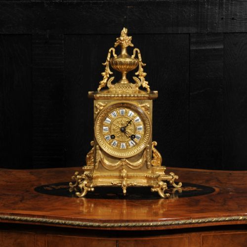 fig 1. A Louis XVI style gilt bronze mantel clock surmounted by a classical urn finial and detailed with drapery swags around a round dial.