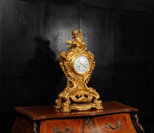 fig 3. An ornate French Rococo ormolu mantel clock decorated with an elaborate cherub finial, acanthus leaf scrolls, and a white enamel dial.
