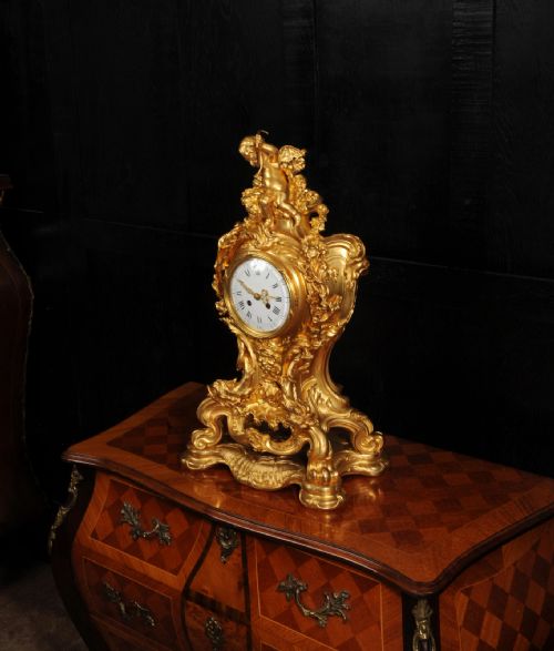 fig 6. An ornate French Rococo ormolu mantel clock decorated with an elaborate cherub finial, acanthus leaf scrolls, and a white enamel dial.