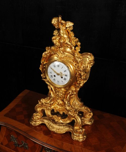 fig 7. An ornate French Rococo ormolu mantel clock decorated with an elaborate cherub finial, acanthus leaf scrolls, and a white enamel dial.
