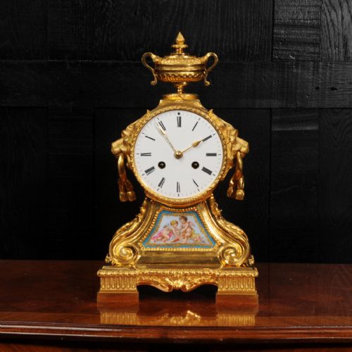 fig 1. A gilded French ormolu drum head mantel clock with an urn finial, lion mask side handles, a white enamel dial, and a hand-painted porcelain panel depicting putti.