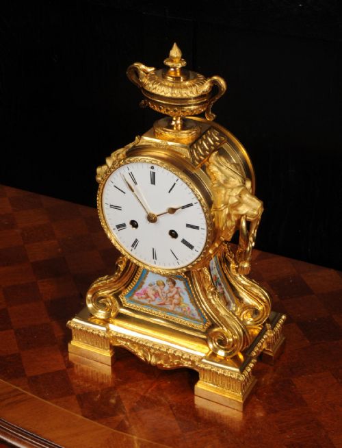 fig 2. A gilded French ormolu drum head mantel clock with an urn finial, lion mask side handles, a white enamel dial, and a hand-painted porcelain panel depicting putti.