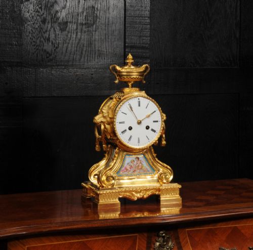 fig 3. A gilded French ormolu drum head mantel clock with an urn finial, lion mask side handles, a white enamel dial, and a hand-painted porcelain panel depicting putti.