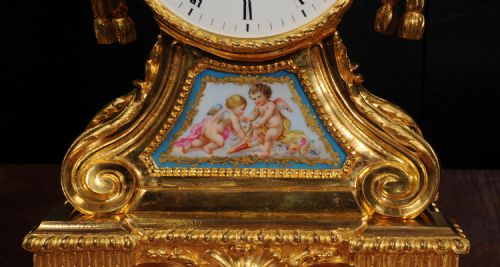 fig 4. A gilded French ormolu drum head mantel clock with an urn finial, lion mask side handles, a white enamel dial, and a hand-painted porcelain panel depicting putti.