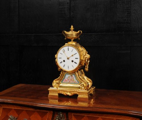 fig 5. A gilded French ormolu drum head mantel clock with an urn finial, lion mask side handles, a white enamel dial, and a hand-painted porcelain panel depicting putti.