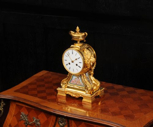 fig 6. A gilded French ormolu drum head mantel clock with an urn finial, lion mask side handles, a white enamel dial, and a hand-painted porcelain panel depicting putti.