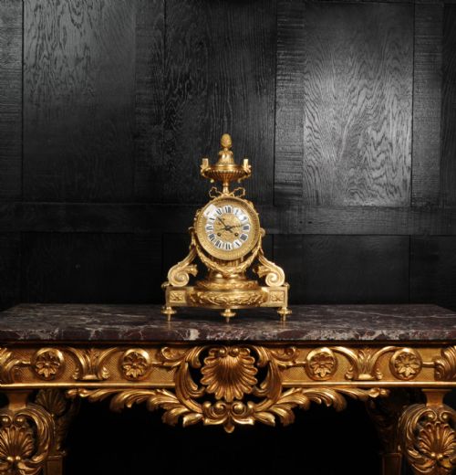 fig 1. A French gilt bronze drum head mantel clock featuring a classical urn finial, laurel wreath garlands, scroll supports, and an enamel cartouche Roman numeral dial.
