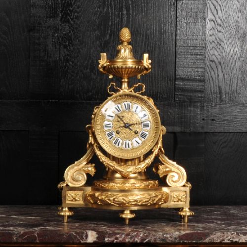 fig 2. A French gilt bronze drum head mantel clock featuring a classical urn finial, laurel wreath garlands, scroll supports, and an enamel cartouche Roman numeral dial.