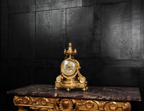 fig 3. A French gilt bronze drum head mantel clock featuring a classical urn finial, laurel wreath garlands, scroll supports, and an enamel cartouche Roman numeral dial.