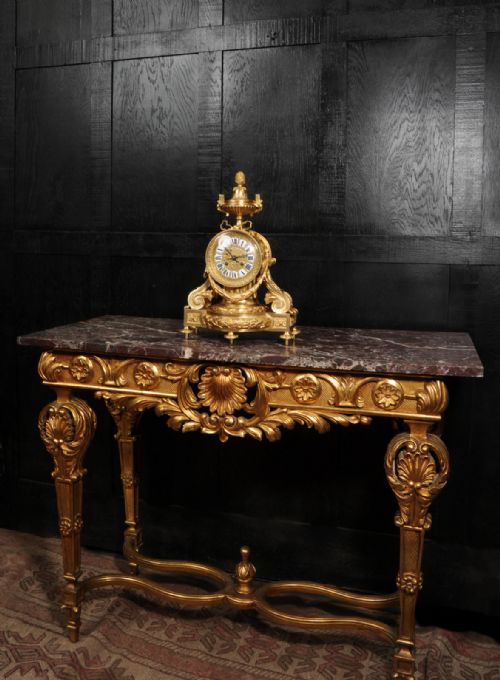 fig 4. A French gilt bronze drum head mantel clock featuring a classical urn finial, laurel wreath garlands, scroll supports, and an enamel cartouche Roman numeral dial.