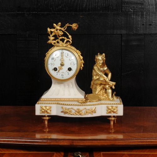fig 1. An antique French mantel clock featuring a white marble base decorated with gilt bronze mounts, an off-centre enamel dial, and a gilded bronze putto figure seated alongside.