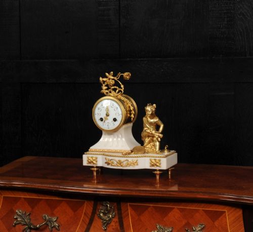 fig 3. An antique French mantel clock featuring a white marble base decorated with gilt bronze mounts, an off-centre enamel dial, and a gilded bronze putto figure seated alongside.