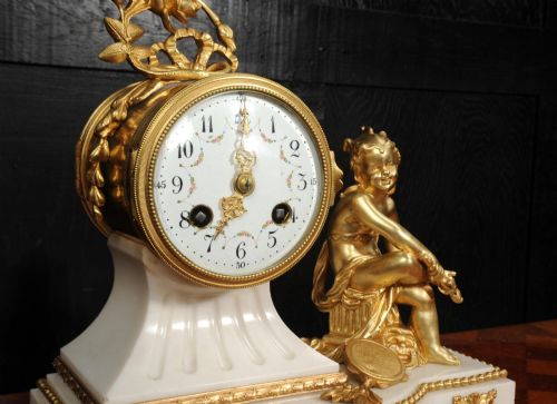 fig 5. An antique French mantel clock featuring a white marble base decorated with gilt bronze mounts, an off-centre enamel dial, and a gilded bronze putto figure seated alongside.